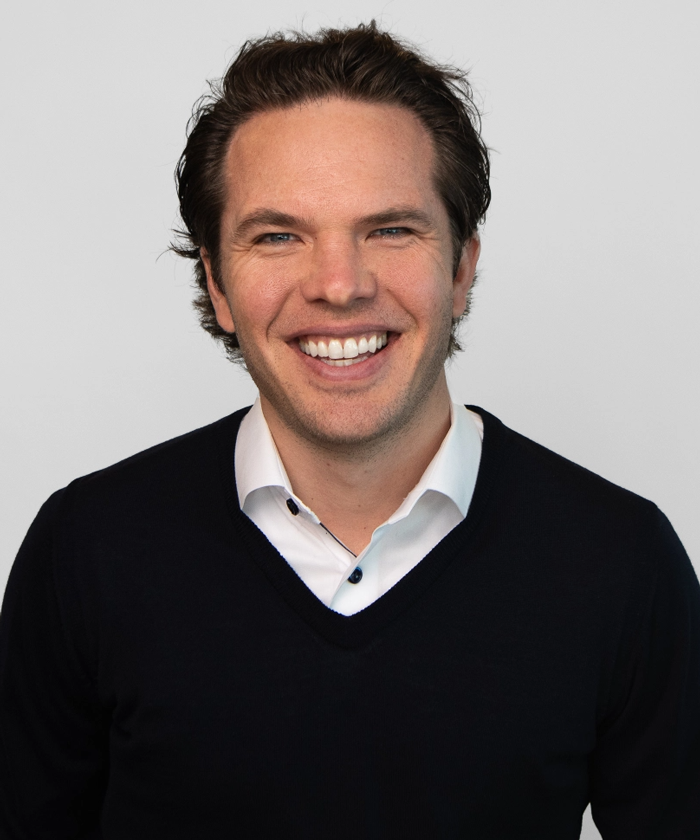 Trommeter Orthodontics | Professional portrait of a smiling man in a black sweater and white collared shirt, standing against a white background.