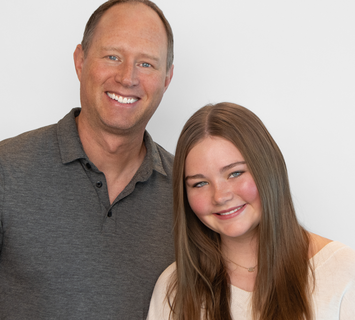 Trommeter Orthodontics | Portrait of a smiling man in a gray polo standing next to a smiling teenage girl with long straight brown hair, wearing a white top.